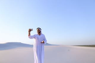 Happy, handsome guy, emirate and tourist, conducts dialogue through Internet with help of device, waves hand and smiles at camera of smartphone, shows beautiful views and sights of large sandy desert outdoors on summer day